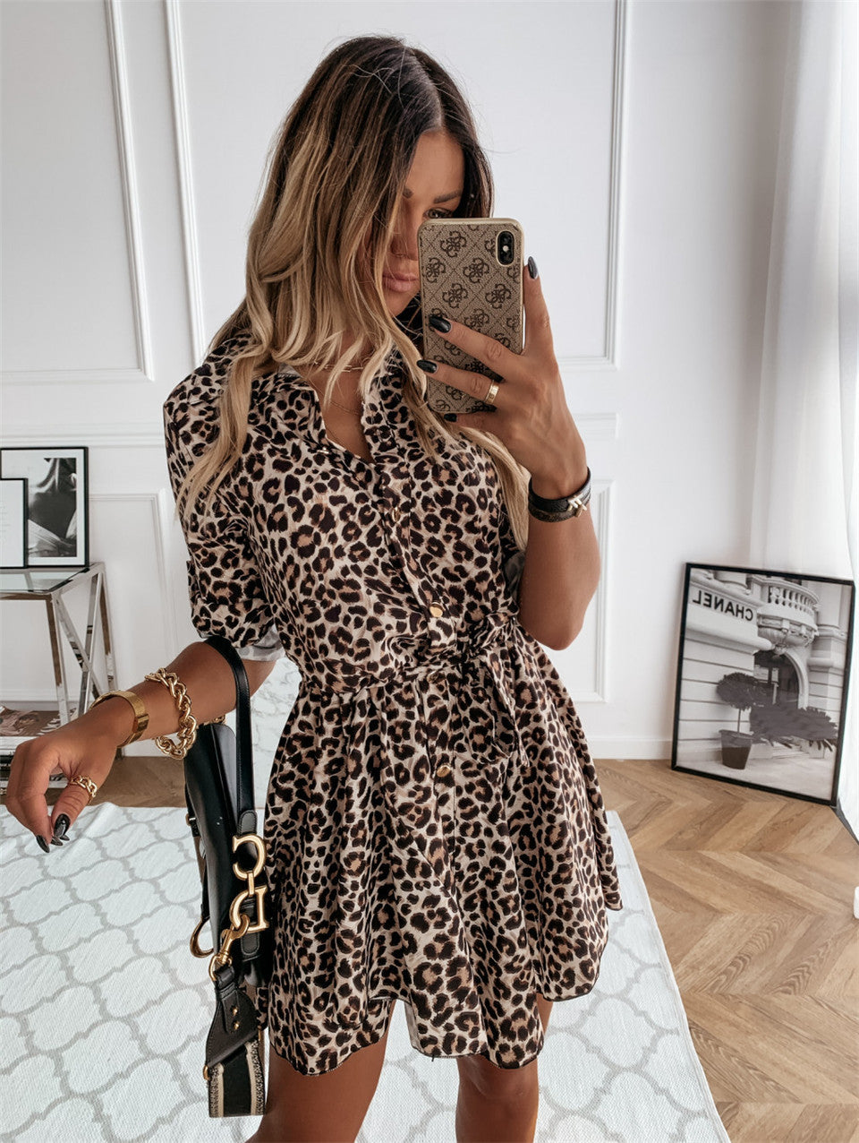 Woman taking a mirror selfie wearing a leopard print dress in a room with a white wall and wooden floor.